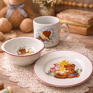 May include: A charming children's tableware set featuring a white bowl, plate, and mug with red trim. Each piece is adorned with whimsical illustrations of a teddy bear. A plush teddy bear and other decorative items are in the background.