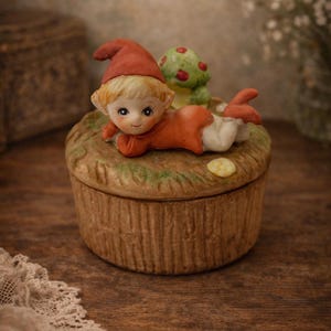 Puede incluir: Una caja de baratijas de cerámica con una figura de elfo con sombrero y atuendo rojos, acostada sobre una tapa con forma de hongo. La caja tiene una apariencia texturizada de madera, con una combinación de colores verde y marrón. También hay un pequeño hongo verde con puntos rojos en la tapa.