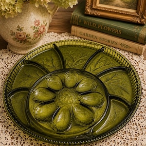 May include: A vintage green glass serving tray with a leaf design. The tray has a central flower-shaped compartment surrounded by six petal-shaped sections. The tray is on a lace doily, with a floral vase and books in the background.
