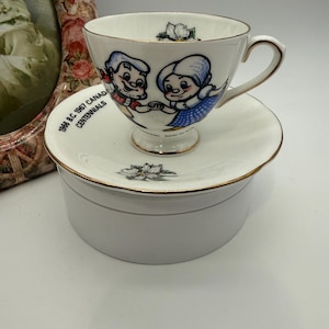 May include: A white teacup and saucer set with gold edging. The teacup features a cartoon couple and a floral design. The saucer has the text "1966 B.C. 1967 CANADA CENTENNIALS". The set is on a white, round box.