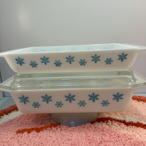 May include: Two white rectangular casserole dishes with teal snowflake designs. One dish has a clear glass lid. The dishes are stacked on top of each other. The dishes are on a white pedestal.