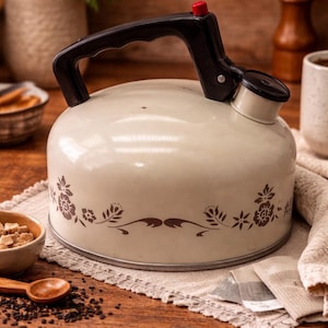 May include: Cream-colored vintage tea kettle with a black handle and spout. The kettle features a floral design in brown and a red knob on the lid. It sits on a wooden surface, suggesting a kitchen or dining setting.