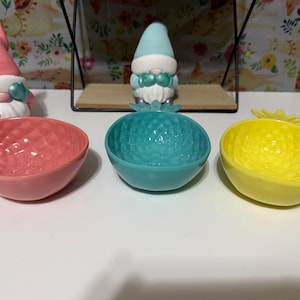 May include: Three small, colourful ceramic bowls in pink, teal, and yellow, each with a textured interior. Decorative gnome figurines are in the background. The bowls are likely for serving snacks or small items.