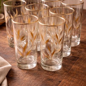 May include: A set of eight clear highball glasses with gold rims and a gold leaf design. The glasses are arranged on a dark wooden surface, suggesting a vintage or retro aesthetic. The glasses are ideal for serving beverages.