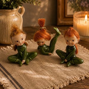 May include: Three ceramic figurines featuring small figures with bright orange hair and dark green outfits. Two figures are sitting in a cross-legged position, while the center figure is lying on its stomach with legs raised in the air.