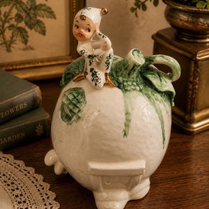 May include: A vintage ceramic figurine of an elf atop a white turnip with green leaves. The elf wears a white outfit with green detailing and a pointed hat. The turnip has a textured surface. The figurine is displayed on a wooden table.