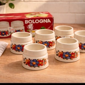 Peut inclure: Ensemble de six coquetiers en céramique vintage avec un motif floral rouge, bleu et orange. Les coquetiers crème ont une forme cylindrique. Une boîte rouge avec l'inscription "BOLOGNA" est visible en arrière-plan.