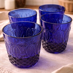 May include: Four cobalt blue drinking glasses. Each glass has a leaf pattern around the sides and a diamond pattern at the base. The glasses are sitting on a light-colored cloth.