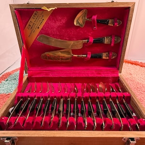 May include: A vintage wooden cutlery set with a red velvet interior. The set includes gold-toned utensils with black handles, including knives, forks, spoons, and serving pieces. The box has a label that reads "JIM'S GEMS".