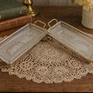 May include: Two rectangular glass trays with gold handles and trim. The trays feature a textured, patterned design and are set on a cream lace doily. The trays are displayed on a wooden surface, with books and a vase of flowers in the background.