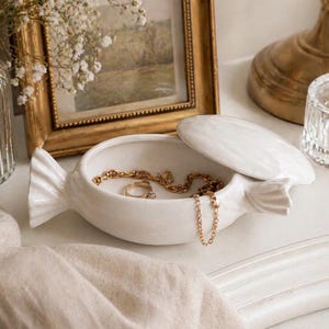 May include: A white ceramic jewellery dish with a lid, shaped like a fish, contains gold necklaces and a ring. The dish is on a white surface, beside a framed picture and a glass perfume bottle.