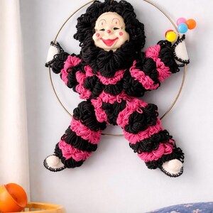 May include: A whimsical clown wall hanging. The clown is made of black and pink yarn, with a smiling face and a black curly wig. It holds a cluster of colorful balloons. The clown is mounted on a gold hoop.