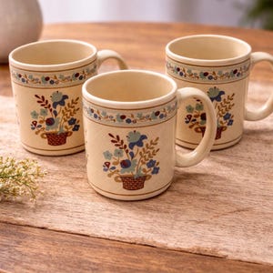 May include: Three cream-colored ceramic mugs with floral designs. Each mug features a basket of blue flowers and a decorative border. The mugs have a handle and are placed on a wooden table.