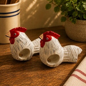 Peut inclure: Deux porte-serviettes en forme de coq en céramique. Les coqs sont blancs avec des crêtes et des becs rouges. Les porte-serviettes sont sur une surface en bois avec un tissu rayé et un porte-ustensiles en céramique en arrière-plan.
