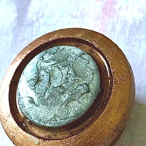 May include: A round, metallic gold-colored object with a central, oval-shaped, textured, teal-colored element. The object has a raised, textured rim and a glossy finish, suggesting a decorative or functional item.