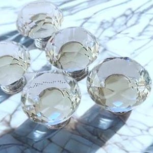 May include: Five clear crystal cabinet knobs with faceted surfaces are arranged on a marble countertop. The knobs have a round shape and a silver-colored base. The lighting creates highlights and shadows on the crystal.