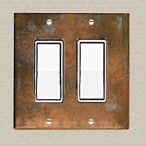 May include: A double light switch wall plate featuring a weathered, oxidized copper finish with a mottled brown and green patina. The rectangular plate contains two white rocker-style switches and is secured by four small, visible metal screws.