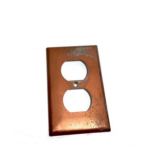 May include: A rectangular copper-toned electrical outlet wall plate featuring two vertical oval openings for a duplex receptacle and a small centered hole for a mounting screw. The metal surface shows signs of wear, patina, and discoloration.