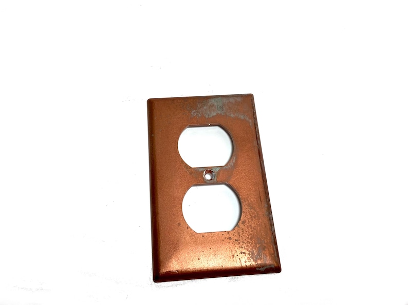 May include: A rectangular copper-toned electrical outlet wall plate featuring two vertical oval openings for a duplex receptacle and a small centered hole for a mounting screw. The metal surface shows signs of wear, patina, and discoloration.
