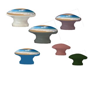 Op de afbeelding: Een verzameling decoratieve kastknoppen in verschillende kleuren, waaronder wit, grijs, blauw, roze en groen. Elke knop heeft een strandsc&egrave;ne met een blauwe lucht, witte wolken en een zandstrand. De knoppen zijn paddenstoelvormig.