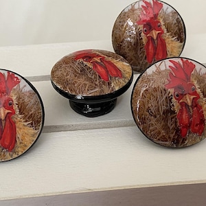 May include: Four decorative cabinet knobs with a rooster design. The knobs have a glossy finish and a black base. The rooster image features a red comb and wattle, with a brown and tan background.
