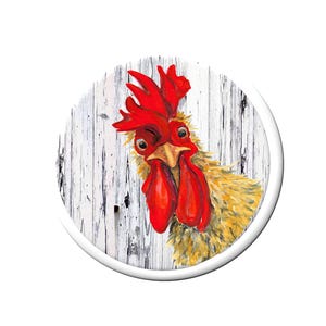 May include: A circular decorative plaque featuring a detailed painting of a cockerel with bright red wattles and a comb, set against a background of weathered white vertical wooden planks. The cockerel has yellow and brown feathers.