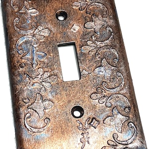 May include: A decorative light switch cover with an aged copper finish. The cover features embossed floral and scrollwork designs with hints of blue and silver. It has a rectangular opening for the switch and two screw holes.