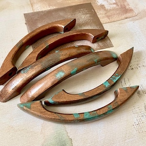 May include: Six copper-coloured cabinet pulls with a curved design. The pulls have a distressed finish with hints of turquoise-green patina. The hardware is arranged on a light-coloured surface with a piece of sandpaper in the background.
