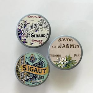 May include: Three vintage-style round tins with labels. One tin reads "Parfume aux Violettes de J. Giraud Grasse Paris" with floral accents. Another says "Savon au Jasmin Paris" with jasmine flowers. The third tin reads "Sigaut Paris".