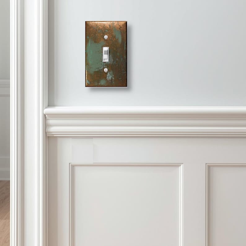 May include: A rectangular light switch cover plate featuring a weathered, oxidized copper finish with mottled patches of metallic bronze and teal green. The plate includes a central toggle switch and two silver-toned mounting screws at the top and bottom.