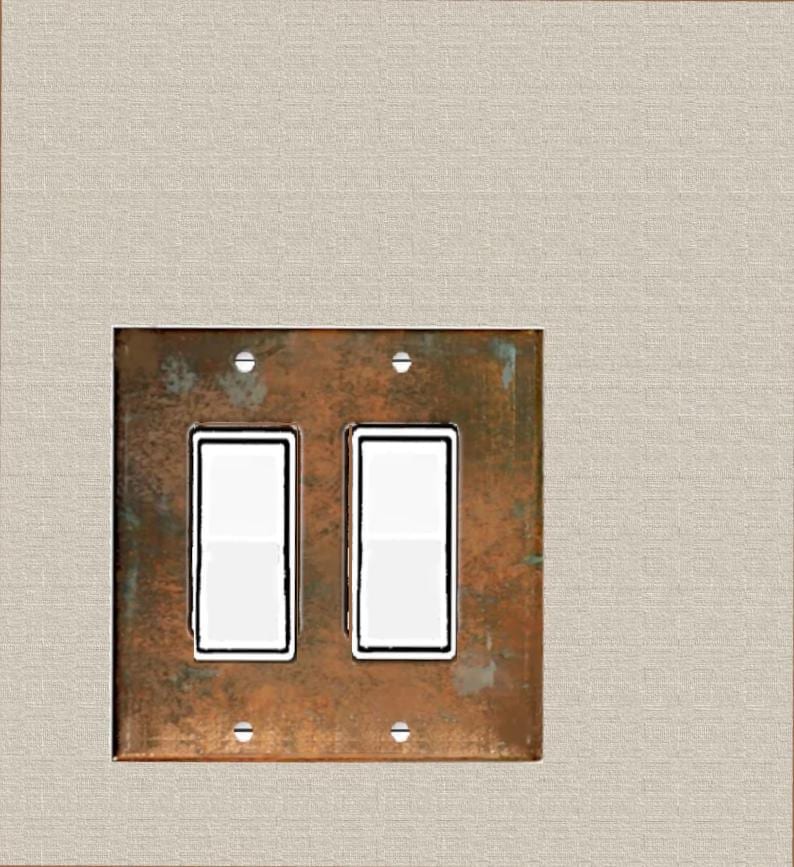 May include: A double light switch wall plate featuring a weathered, oxidized copper finish with a mottled brown and green patina. The rectangular plate contains two white rocker-style switches and is secured by four small, visible metal screws.