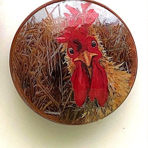May include: A round, wooden button featuring a close-up of a rooster's head. The rooster has bright red comb and wattles, with a yellow beak and a detailed, expressive face. The background shows a field of hay.