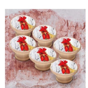 Peut inclure: Un ensemble de six boutons de placard ronds en bois, chacun présentant une illustration imprimée d'une tête de coq avec une crête et des barbillons rouge vif sur un fond de planches de bois verticales blanches. Base en bois clair naturel.