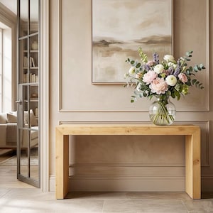 May include: A light-colored wooden console table with a clear glass vase filled with flowers. The flowers are a mix of pink, white, and purple blooms with green foliage. A landscape painting hangs above the table.