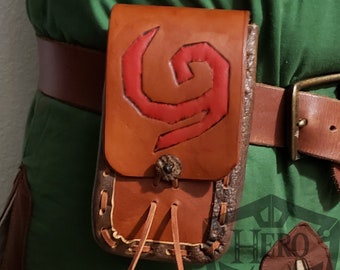 Puede incluir: Una bolsa de cuero marrón con un emblema rojo, sujeta a un cinturón marrón. La bolsa tiene un cierre de solapa y un nudo decorativo. El fondo muestra tela verde y un cinturón marrón.