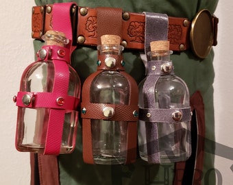 May include: Three glass bottles with cork stoppers and leather straps are attached to a brown leather belt with a metal buckle. The bottles are clear glass and have a brown, pink, and purple leather strap.