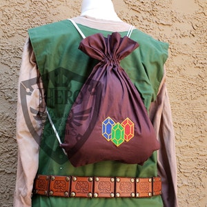 May include: A brown drawstring bag with a white cord and a green, red, and blue gem design. The bag is worn as a backpack over a green tunic with a brown leather belt.