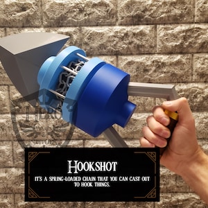 May include: A blue and grey hookshot prop with a handle, designed to cast out a spring-loaded chain. The prop has a blue cylindrical body, a grey handle, and a grey blade-like structure. The text "HOOKSHOT" is displayed on a black background.