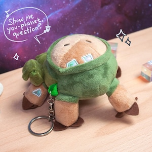 May include: A plush keychain featuring a tan creature with a green hooded garment, brown hooves, and iridescent accents. A speech bubble above the plush contains the text "Show me you-planet, question?" with musical notes. A metal ring is attached.