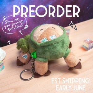 Peut inclure: Peluche en forme de créature avec une veste à capuche verte, des pieds marron et un visage beige. Le texte "PREORDER" est en haut, et une bulle de dialogue dit "Show me you-planet, question?". Le texte "EST SHIPPING: EARLY JUNE" est en bas.