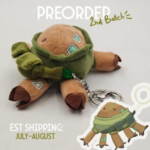 May include: A brown and green plush creature with four legs, a rounded body, and light blue square accents. It features a green fabric strap attached to a silver metal keychain. The text PREORDER 2nd Batch is visible at the top of the image.
