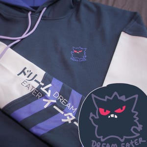 Puede incluir: Una sudadera con capucha azul oscuro con rayas blancas y moradas en las mangas. La sudadera tiene un pequeño gráfico de una criatura morada con ojos rojos y el texto "Dream Eater" en la parte delantera. Las mangas tienen el texto "Dream Eater" en japonés e inglés.
