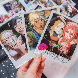 Può includere: Una mano tiene tre stampe in stile polaroid con illustrazioni colorate di personaggi in stile anime. Le stampe mostrano vari personaggi con caratteristiche uniche, tra cui colori di capelli vivaci e design degli occhi. Il testo "Beautiful Night, Love" è scritto su una stampa.