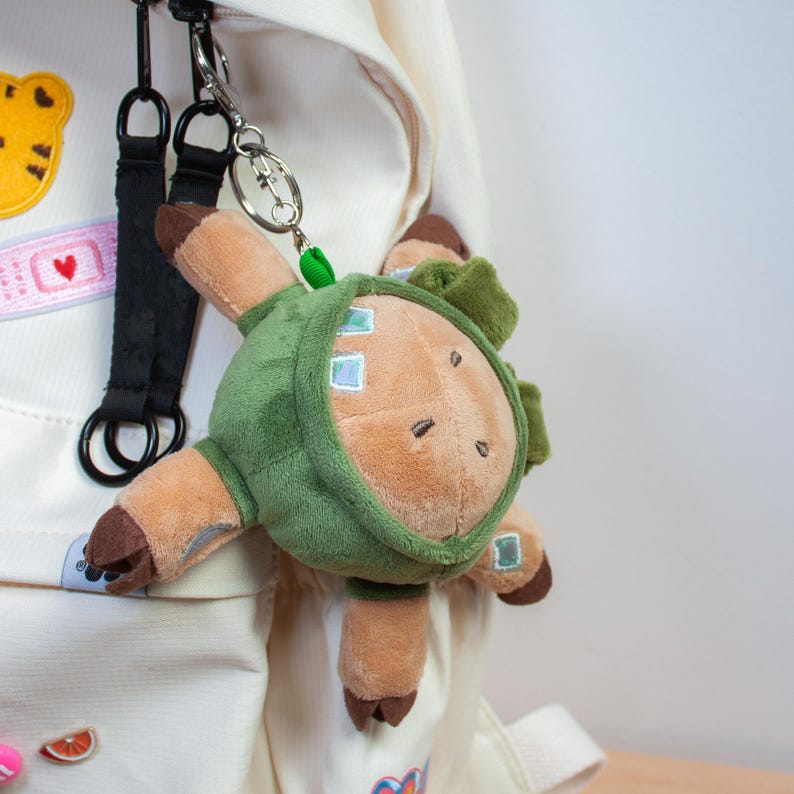 May include: A plush keychain featuring a round, tan creature wearing a dark green hooded cloak. The creature has small brown hooves, simple embroidered eyes, and a small green leaf detail on the hood. It is attached to a silver metal keyring.