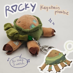 May include: A brown and green plush keychain featuring a creature with four limbs, dark brown hooves, and light blue square accents. The plush has a metal lobster clasp attached. Text on the image reads: ROCKY Keychain plushie and You sleep. I watch.