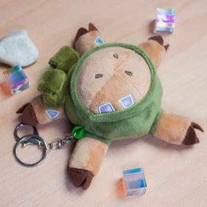 May include: A plush keychain featuring a tan creature with dark brown hooves, wearing a green hooded garment. The creature has light blue square accents on its body and a green fabric detail on its back. A silver metal keyring is attached to the side.