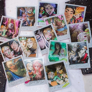 May include: A collection of artistic polaroid-style prints featuring colorful illustrations of characters with various expressions and styles. Some prints include handwritten text like "Rita's stain or sonth?" and "You got this Meli!"