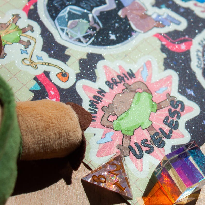 May include: A glittery sticker featuring a brown cartoon figure in a green shirt with the text HUMAN BRAIN USELESS, positioned next to a plush brown fabric edge and a multi-faceted iridescent crystal die on a dark background with grid lines.