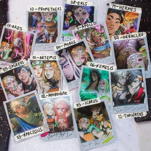May include: A collection of 14 Polaroid-style art prints featuring illustrations of mythological figures. Each print has a unique character design and color palette, with handwritten text and numbers. The prints are arranged on a light-colored surface.
