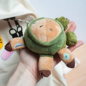 May include: A plush keychain featuring a tan creature wearing a dark olive green hooded garment. The creature has brown hooves, small embroidered eyes, and iridescent blue diamond-shaped accents on its limbs. It is attached to a metal ring.